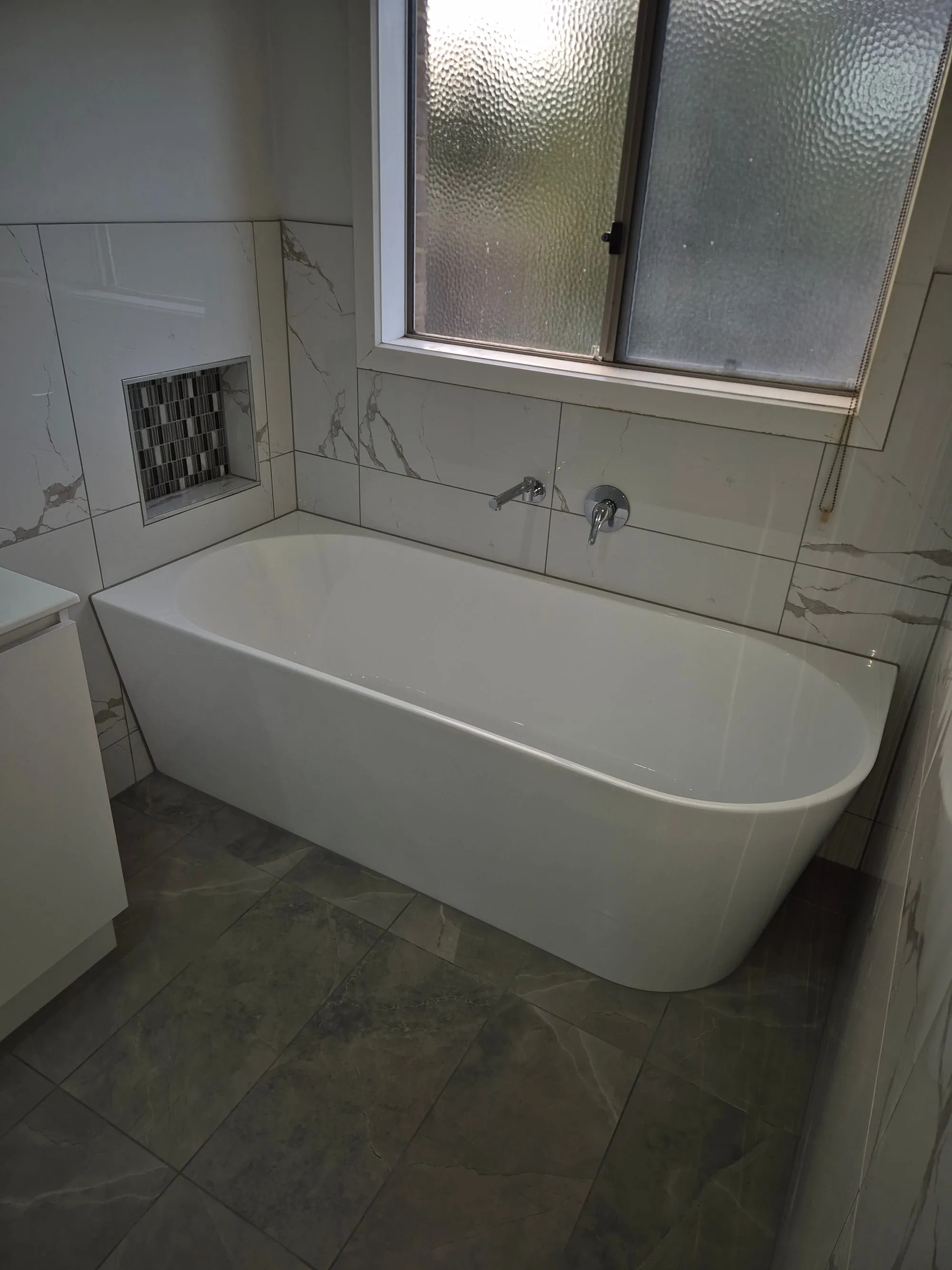Built-in bath below a window, with a mosaic niche in the marble-look tile
